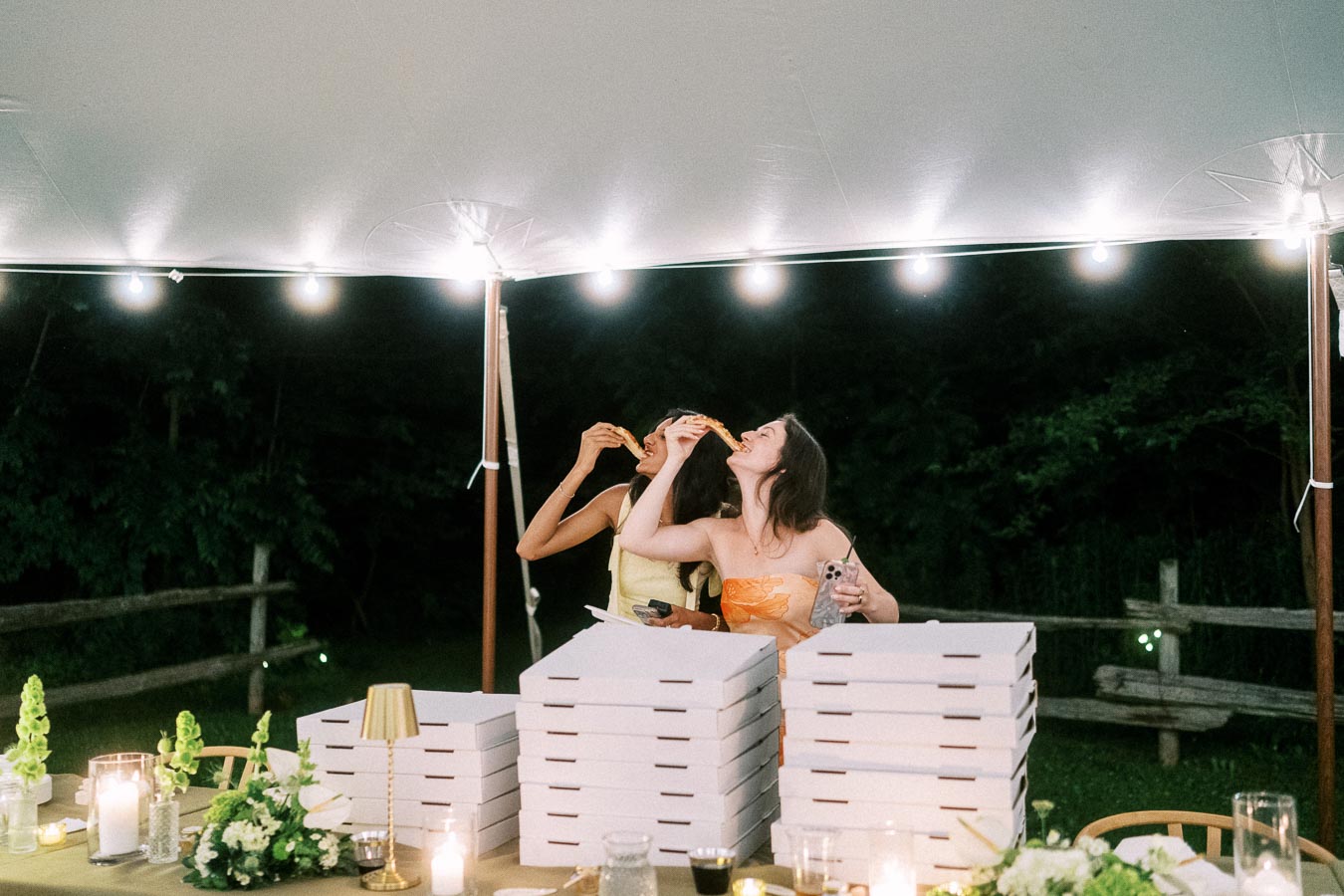 Two women eating pizza under a lit wedding reception tent on a private farm estate near Chatham-Kent, Ontario, Canada.