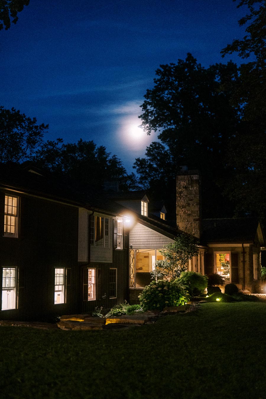 Moonlit private estate farmhouse exterior at night in Chatham-Kent, Ontario, with illuminated windows, garden lights, lawn and trees for an outdoor wedding setting.
