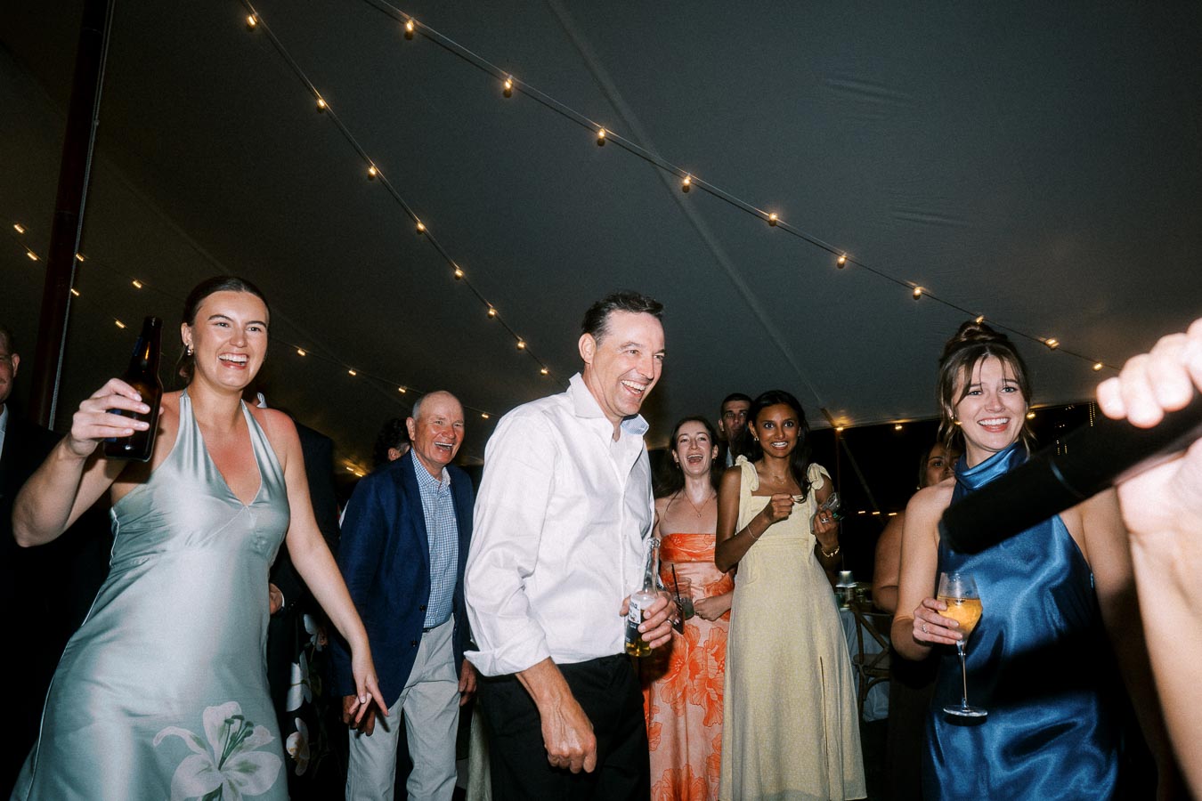 Wedding guests dancing and holding drinks under string lights inside a tent at a private farm estate near Chatham-Kent, Ontario, Canada.