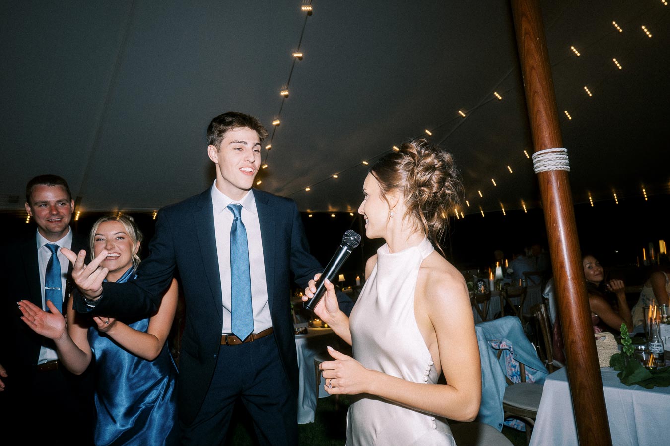 Bride holds a microphone beside smiling wedding guests during a tented reception near Chatham-Kent, Ontario.