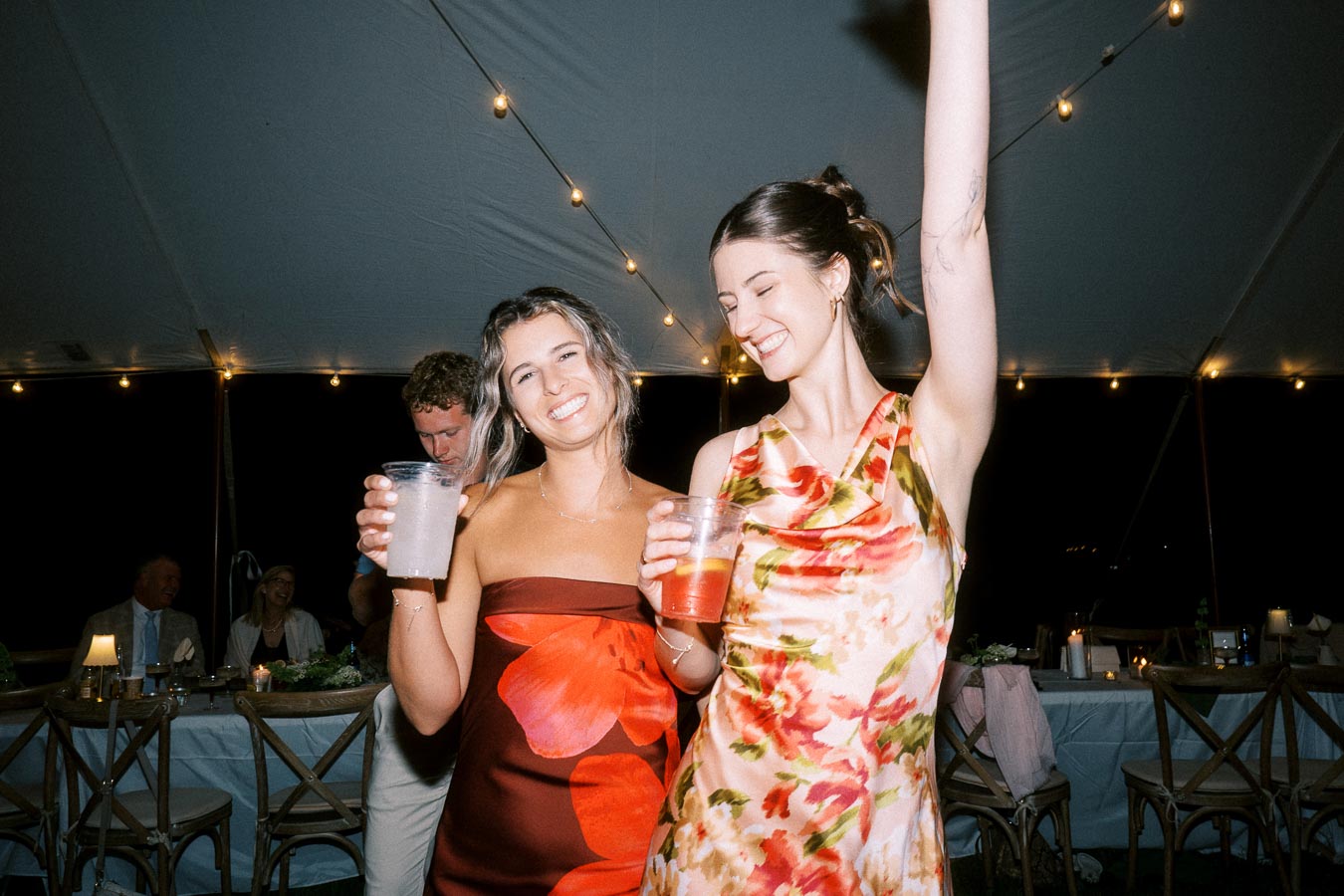 Two women dance with drinks under a wedding reception tent with string lights at night near Chatham-Kent, Ontario, Canada.