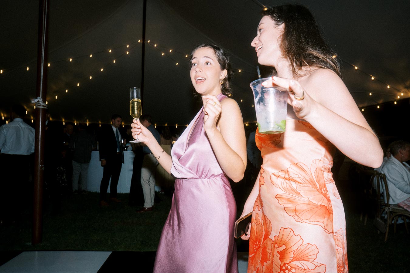 Two women in colorful dresses dance and hold drinks under string lights inside a wedding tent at night near Chatham-Kent, Ontario, Canada.