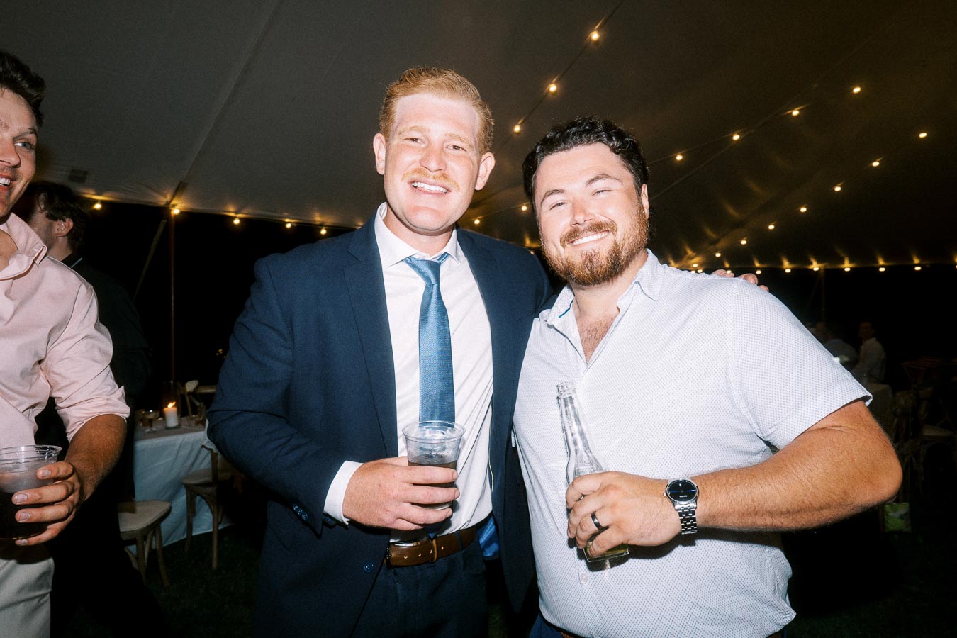 Two smiling male wedding guests holding drinks inside a lit reception tent at a private estate wedding near Chatham-Kent, Ontario, Canada.