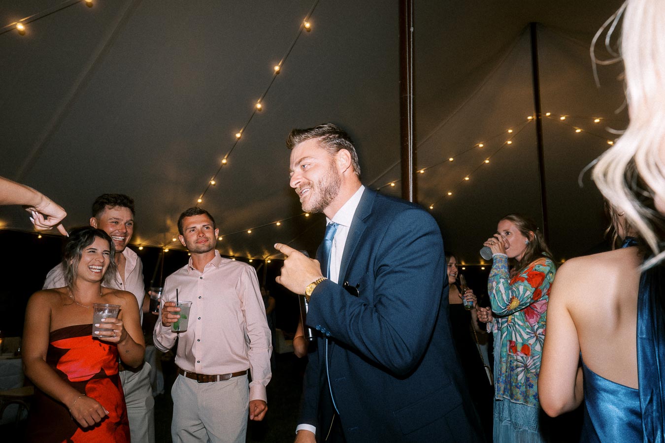 Wedding guests laughing and talking under a tent with string lights at an outdoor private estate reception near Chatham-Kent, Ontario, Canada.
