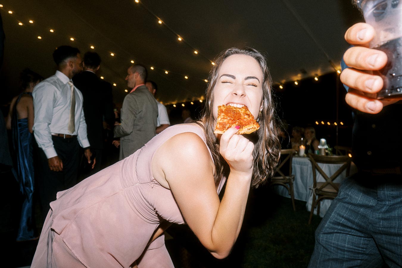 Woman in a pink dress eating pizza under string lights during a tented outdoor wedding reception near Chatham-Kent, Ontario, Canada.
