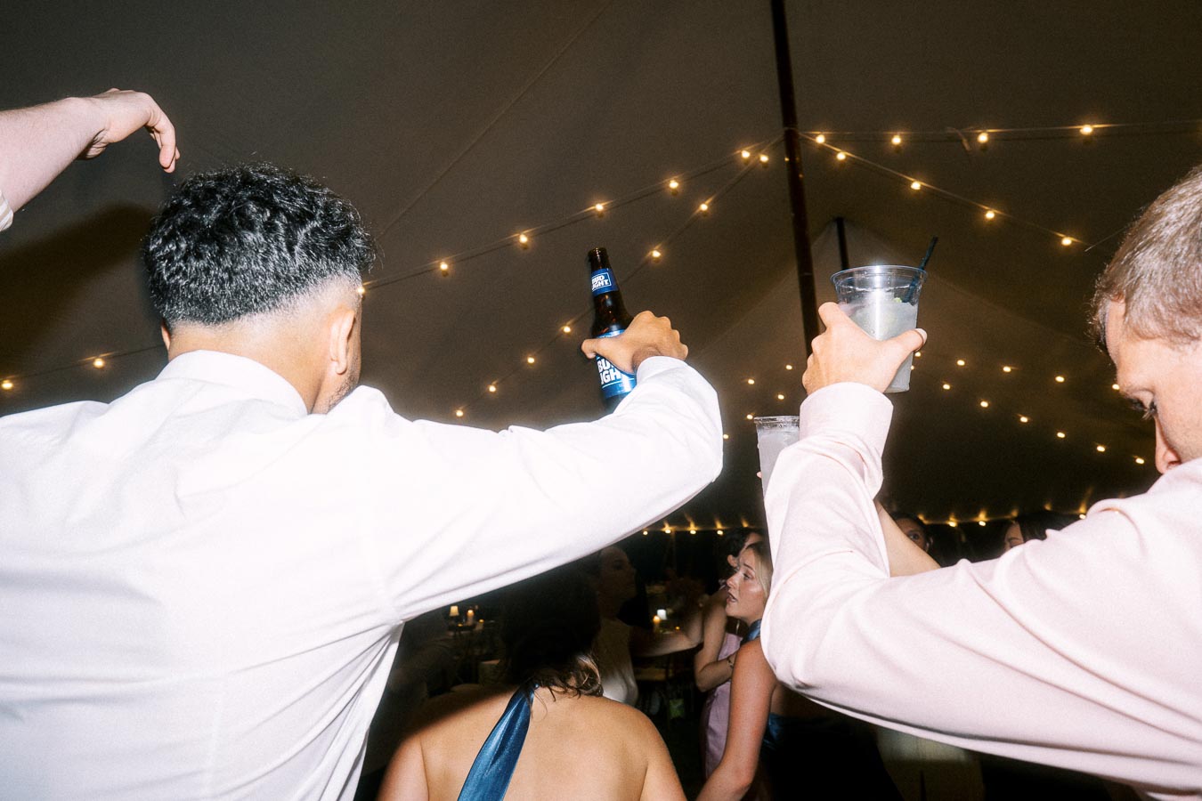 Wedding guests dancing and raising drinks under string lights inside a tent during a private estate farm wedding near Chatham-Kent, Ontario, Canada.