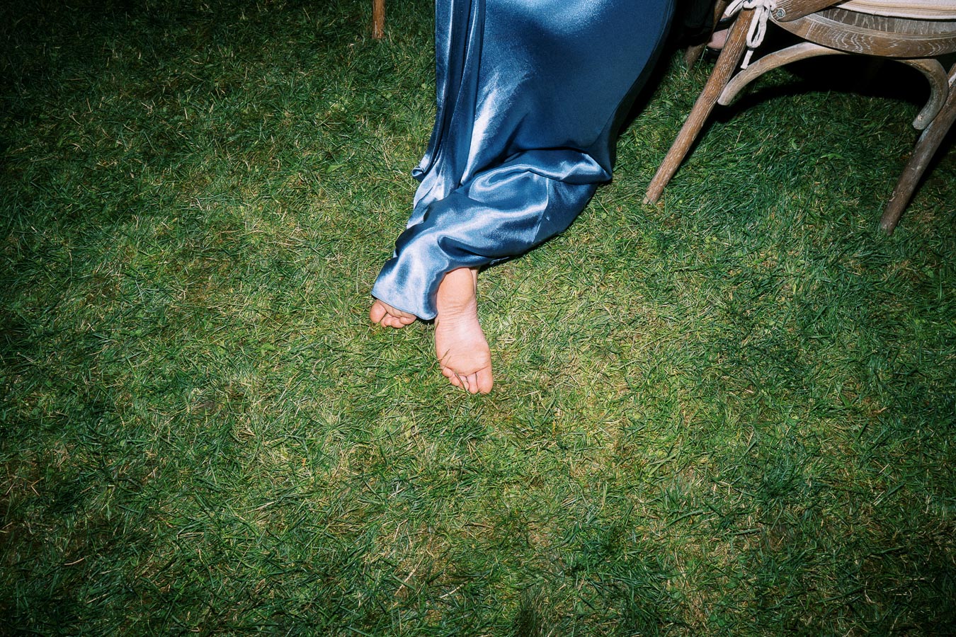 Bare feet under a blue satin dress resting on grass beside a wooden chair at an outdoor farm wedding near Chatham-Kent, Ontario, Canada.
