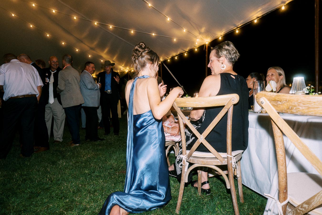 Outdoor wedding reception under a tent with string lights, guests, wooden chairs, and dinner tables on grass at night near Chatham-Kent, Ontario.