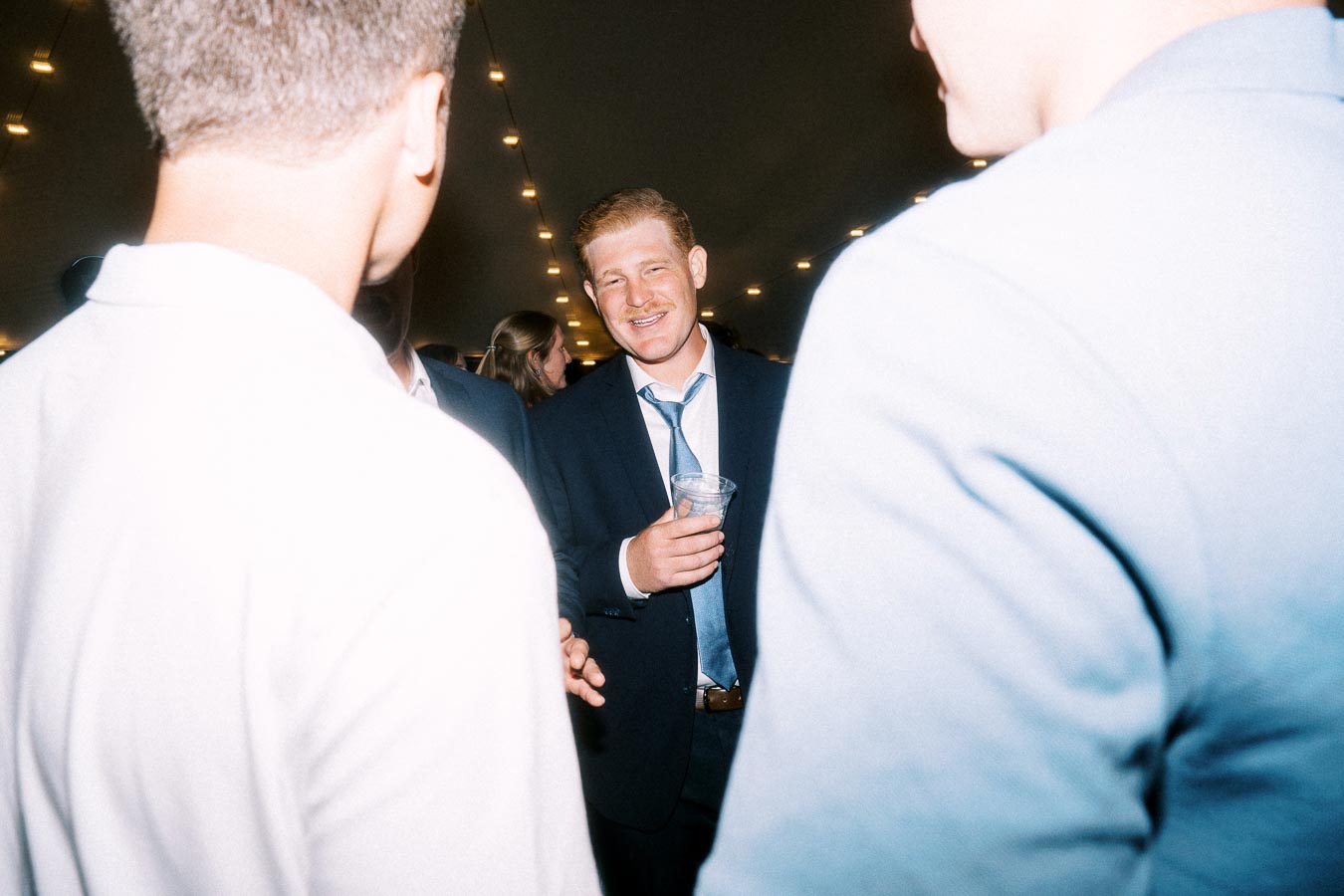 Guests in suits socialize under string lights at a tented wedding reception near Chatham-Kent, Ontario, Canada.