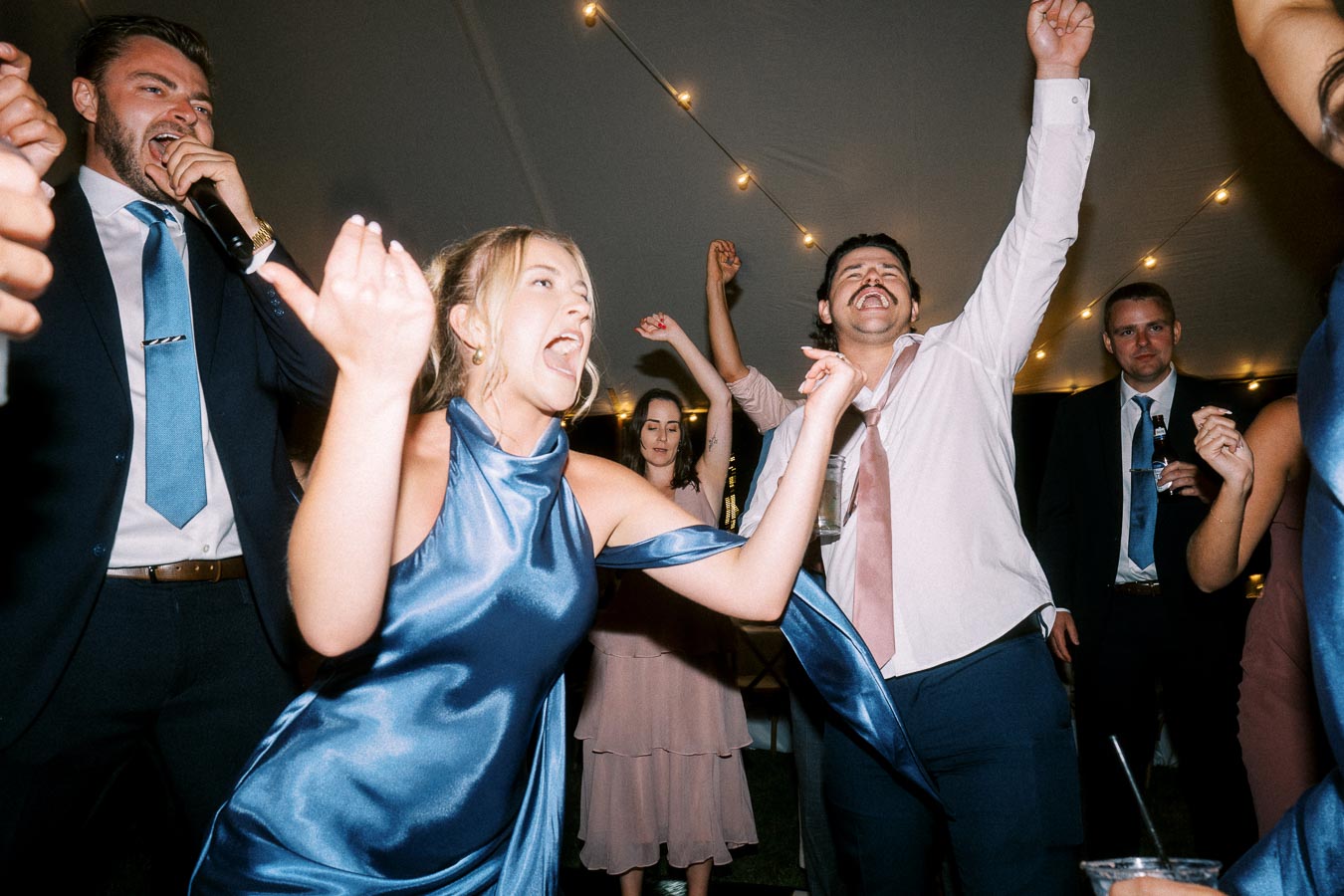Wedding guests dancing and singing inside a tented reception near Chatham-Kent, Ontario, with string lights overhead.