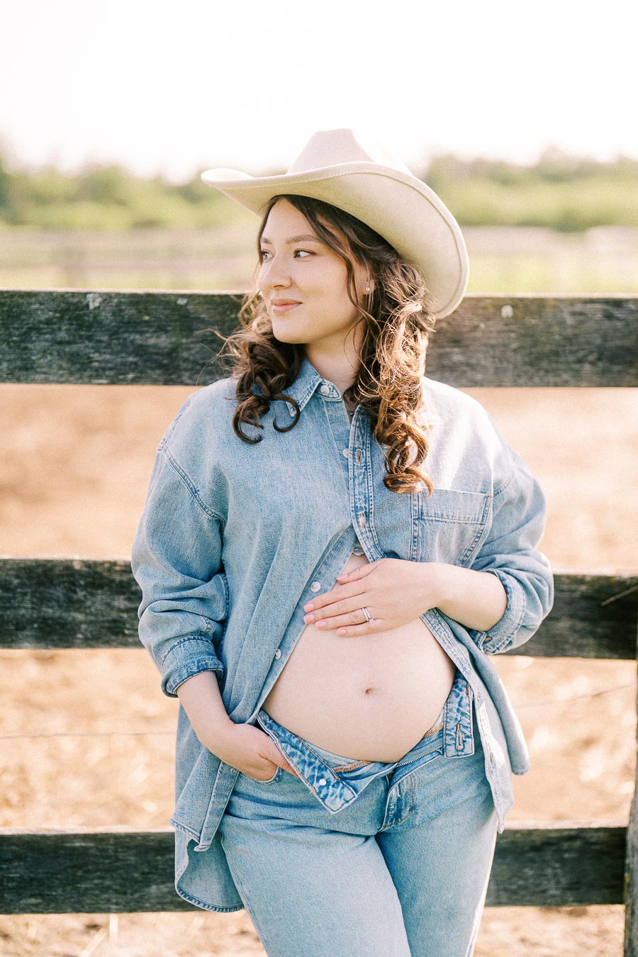 Pregnant woman in denim and cowboy hat by a farm fence