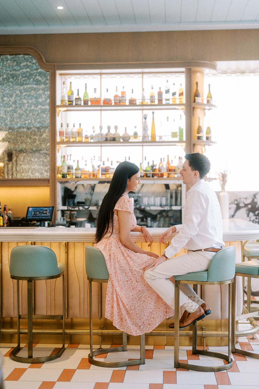 Couple seated at a pastel bar with backlit bottles during their engagement session