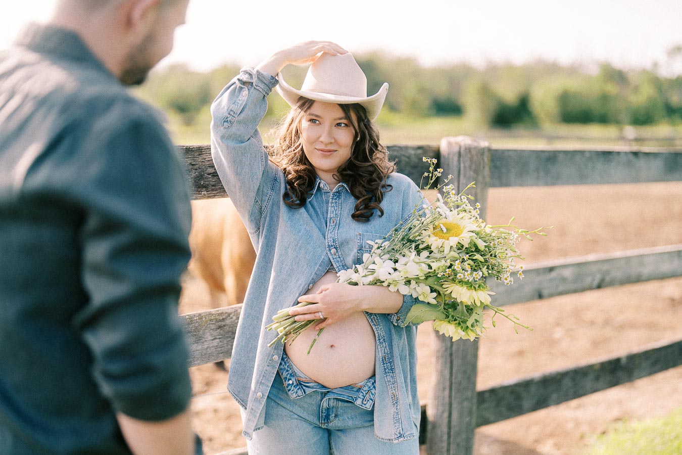 Pregnant woman in cowboy hat holding flowers by a farm fence