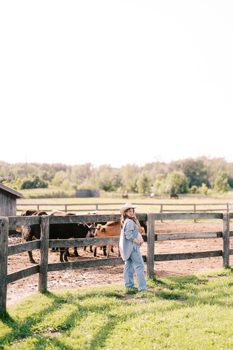 Pregnant woman in cowboy hat by a farm fence with cattle