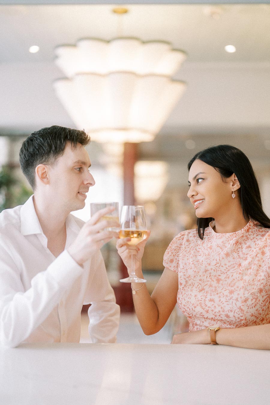 Couple toasting wine glasses indoors beneath a large soft chandelier