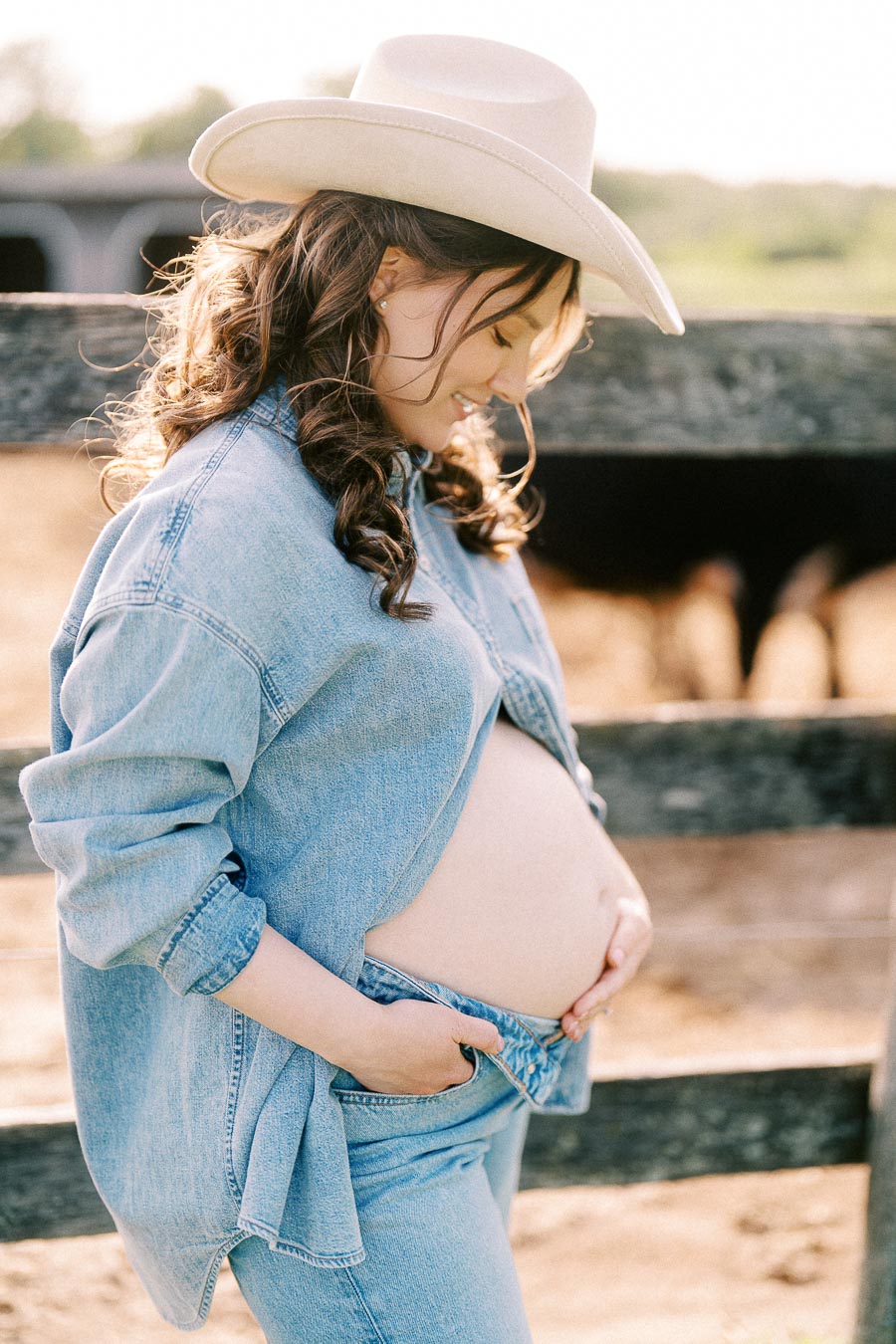 Pregnant woman in denim and cowboy hat by a wooden farm fence