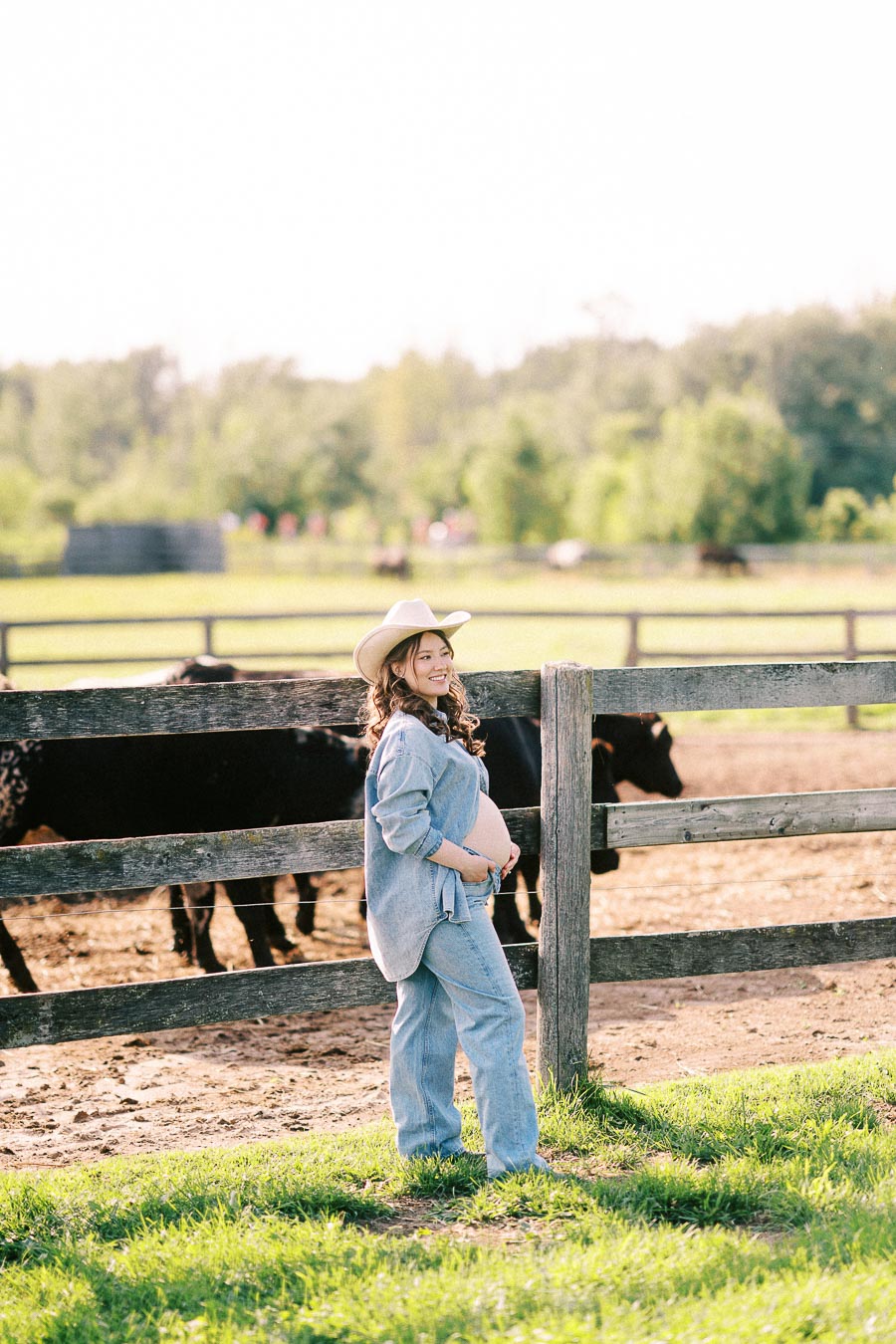 Pregnant woman in cowboy hat standing by a fence with horses behind her