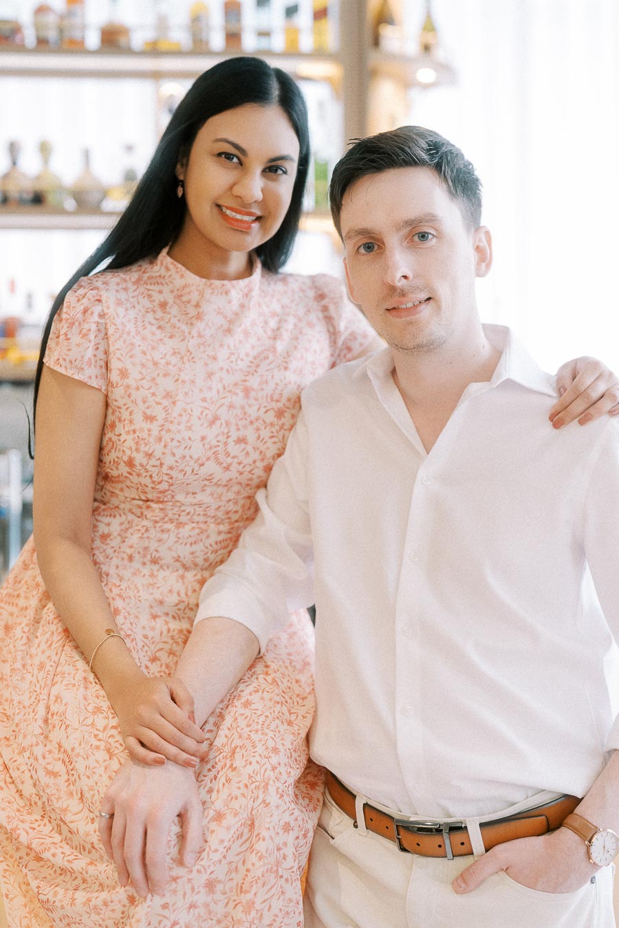 Couple seated together indoors, holding hands beside softly lit shelves