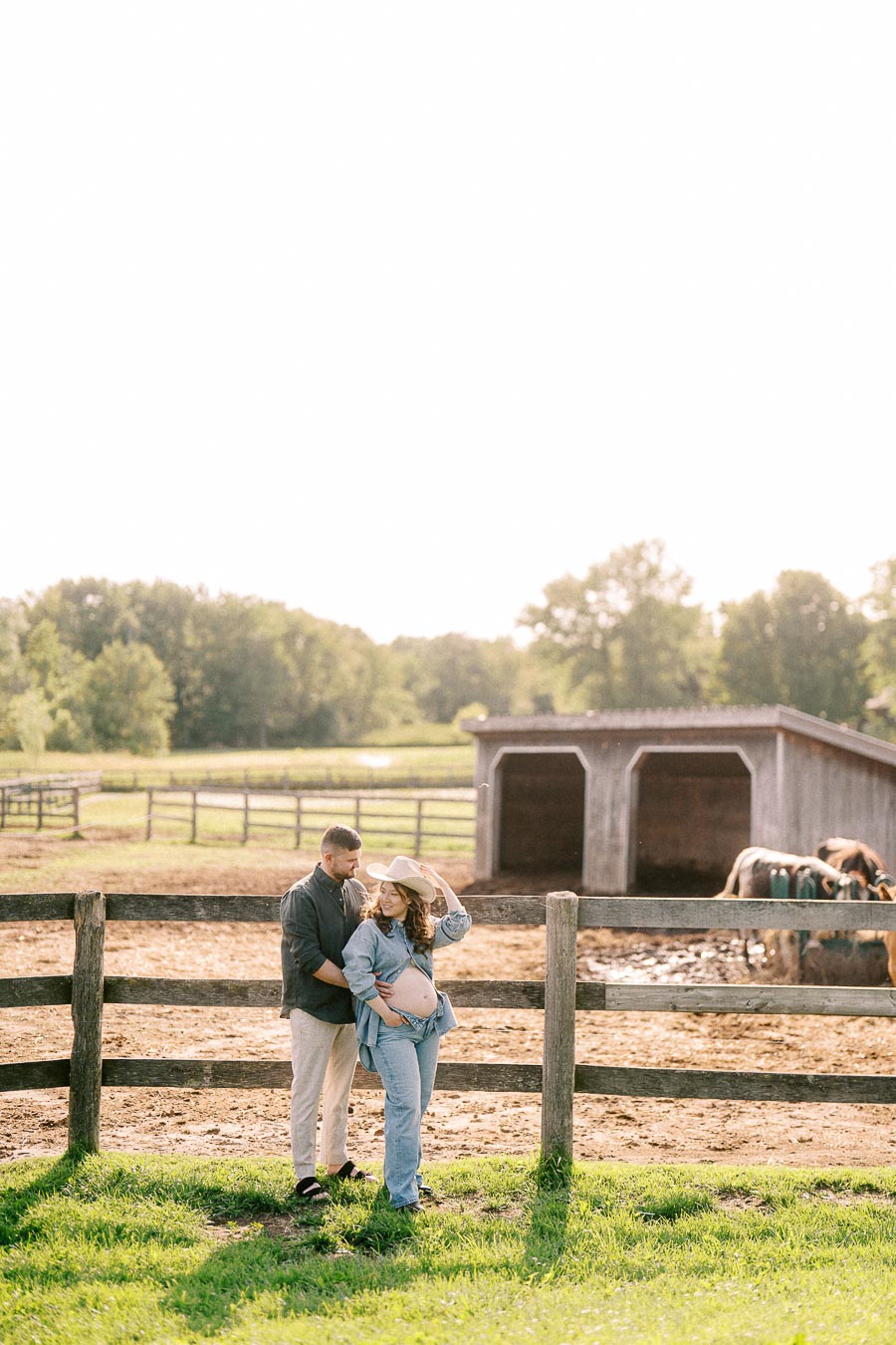 Expectant couple by a paddock fence with horses at The Green Acres