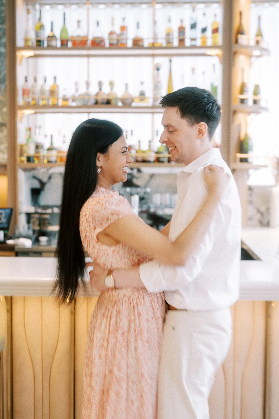 Couple smiling in an embrace beside a bright bar with bottles behind them