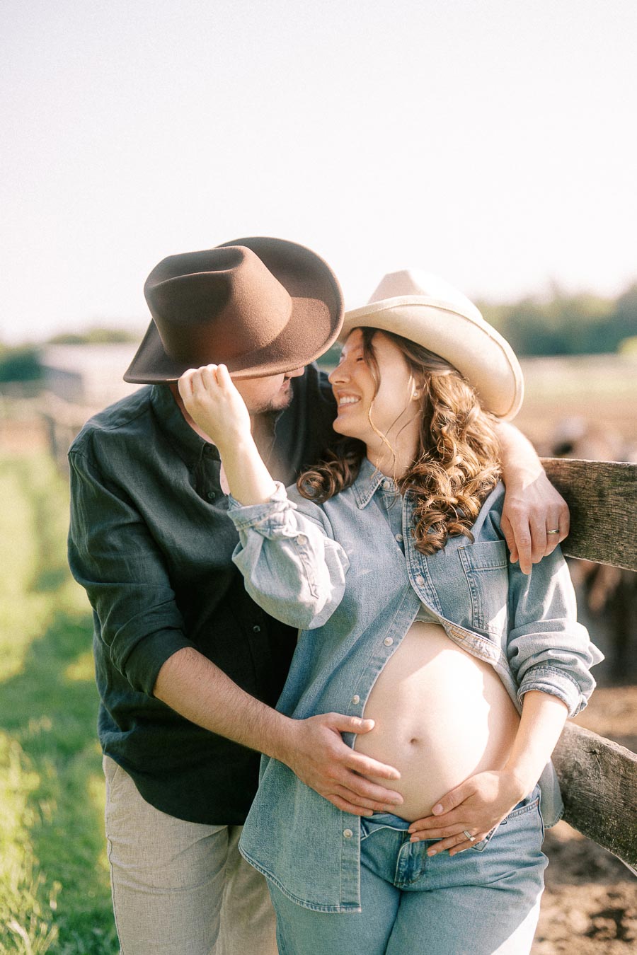 Expectant couple in cowboy hats embracing by a fence at The Green Acres