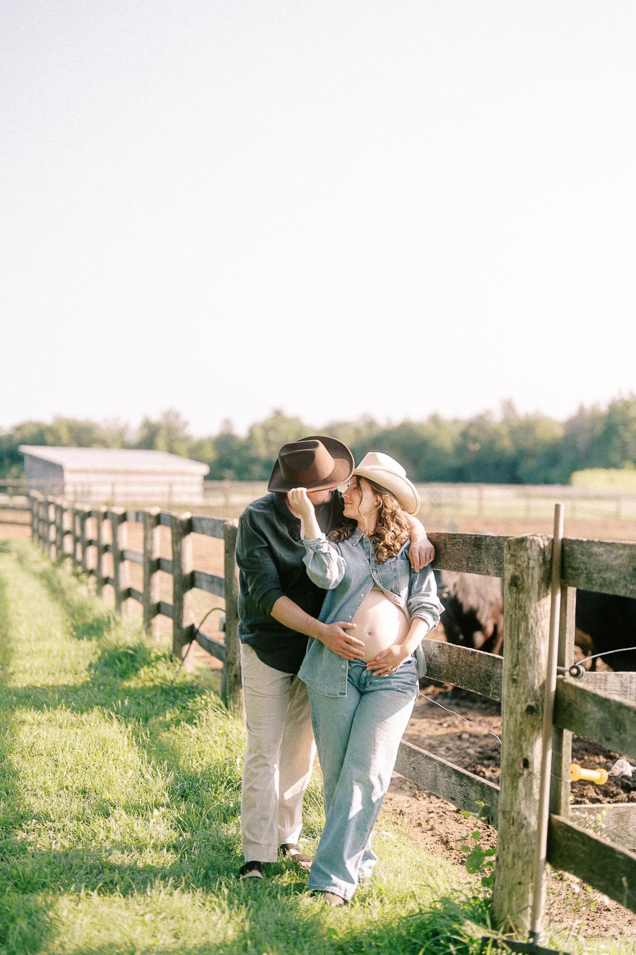Expectant couple in hats embracing by a farm fence at The Green Acres
