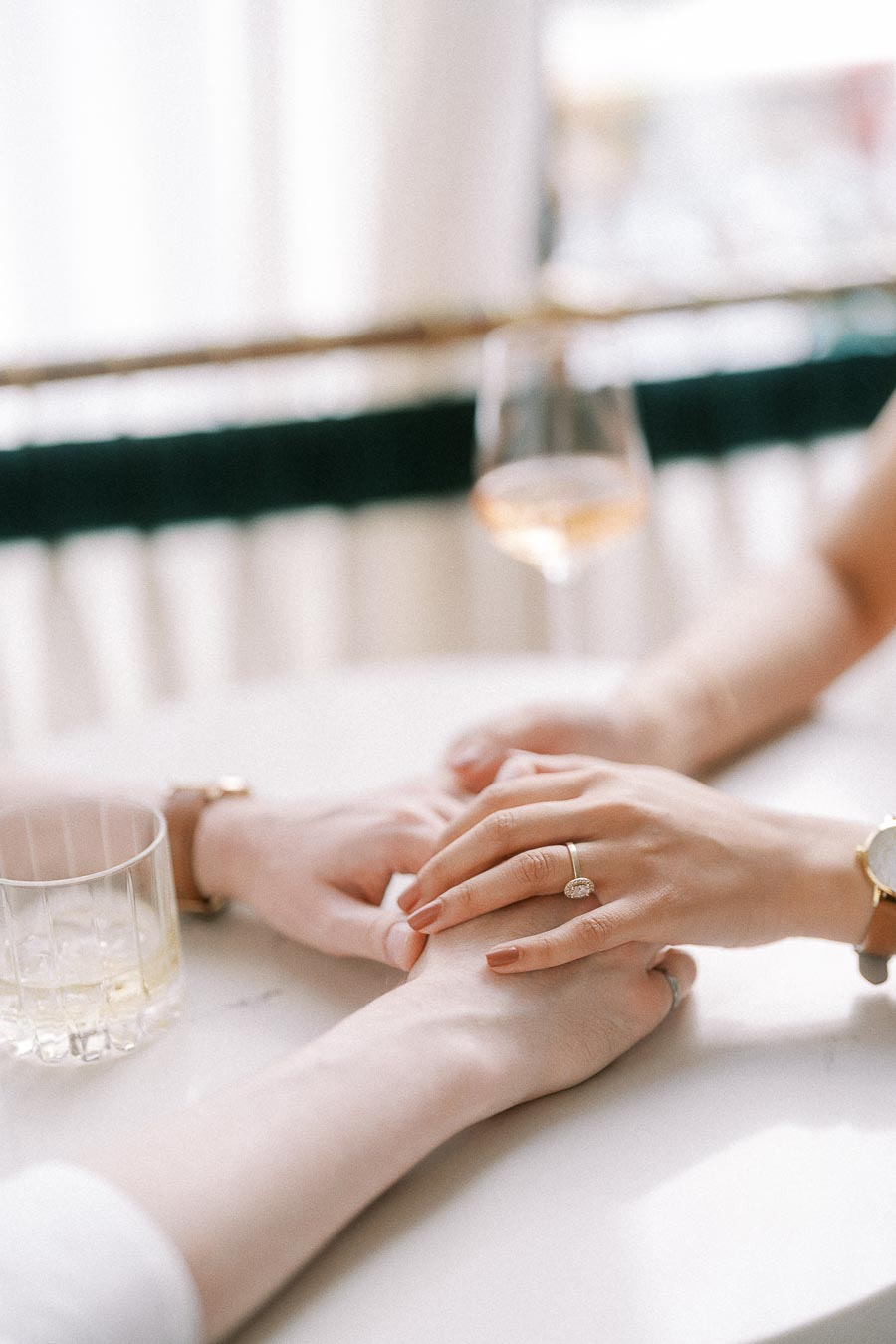 Hands with engagement ring resting together beside drinks at a bright table