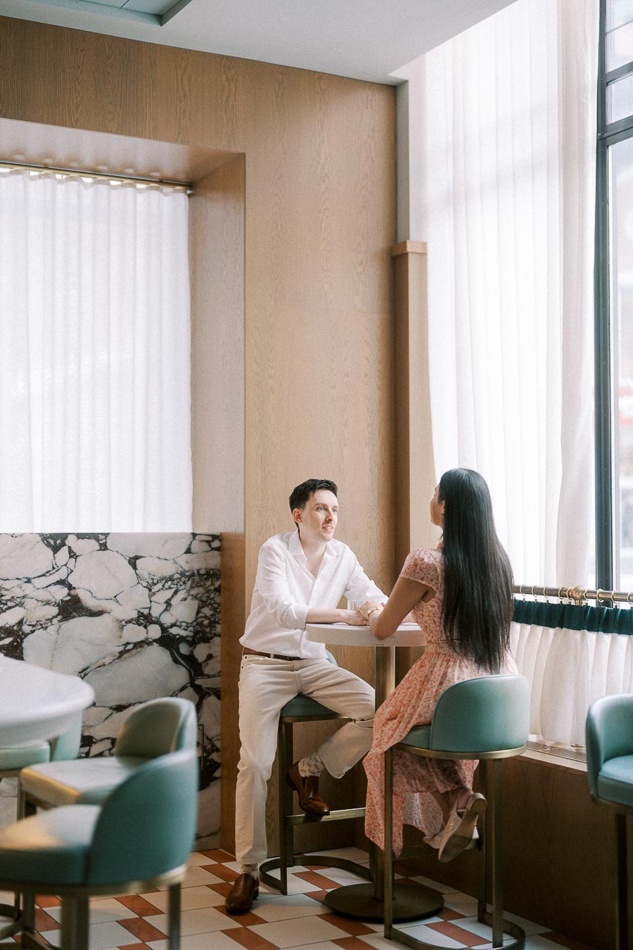 Couple seated at a small café table beside tall windows and white curtains