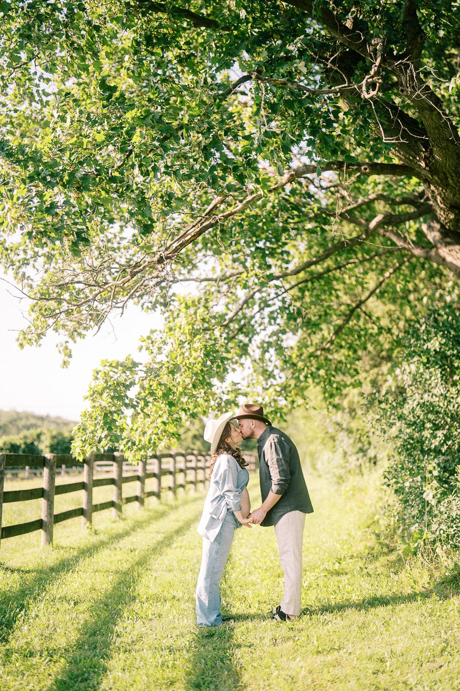 Expectant couple kissing beneath leafy trees beside a farm fence