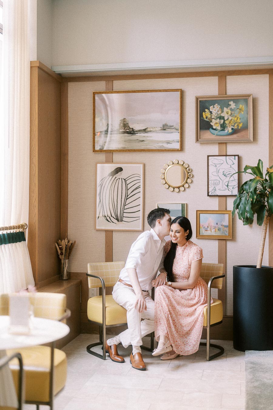Couple seated in yellow chairs, sharing a kiss beside a gallery wall and plant