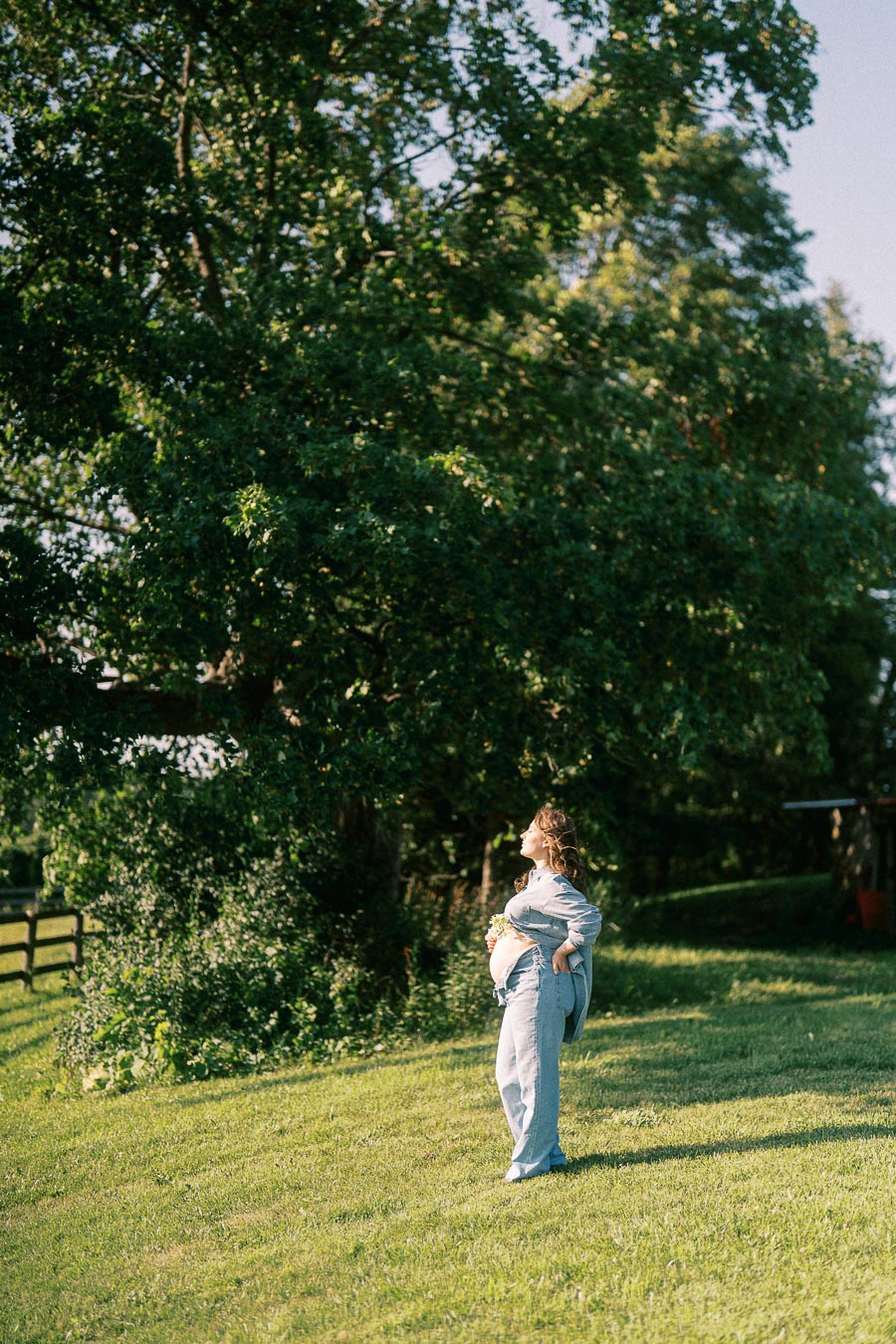 Pregnant woman standing in a grassy field beneath tall trees at The Green Acres