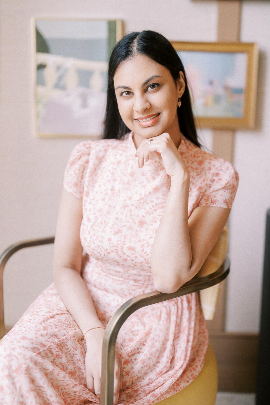 Becky seated indoors in a pink floral dress, smiling with her hand under her chin