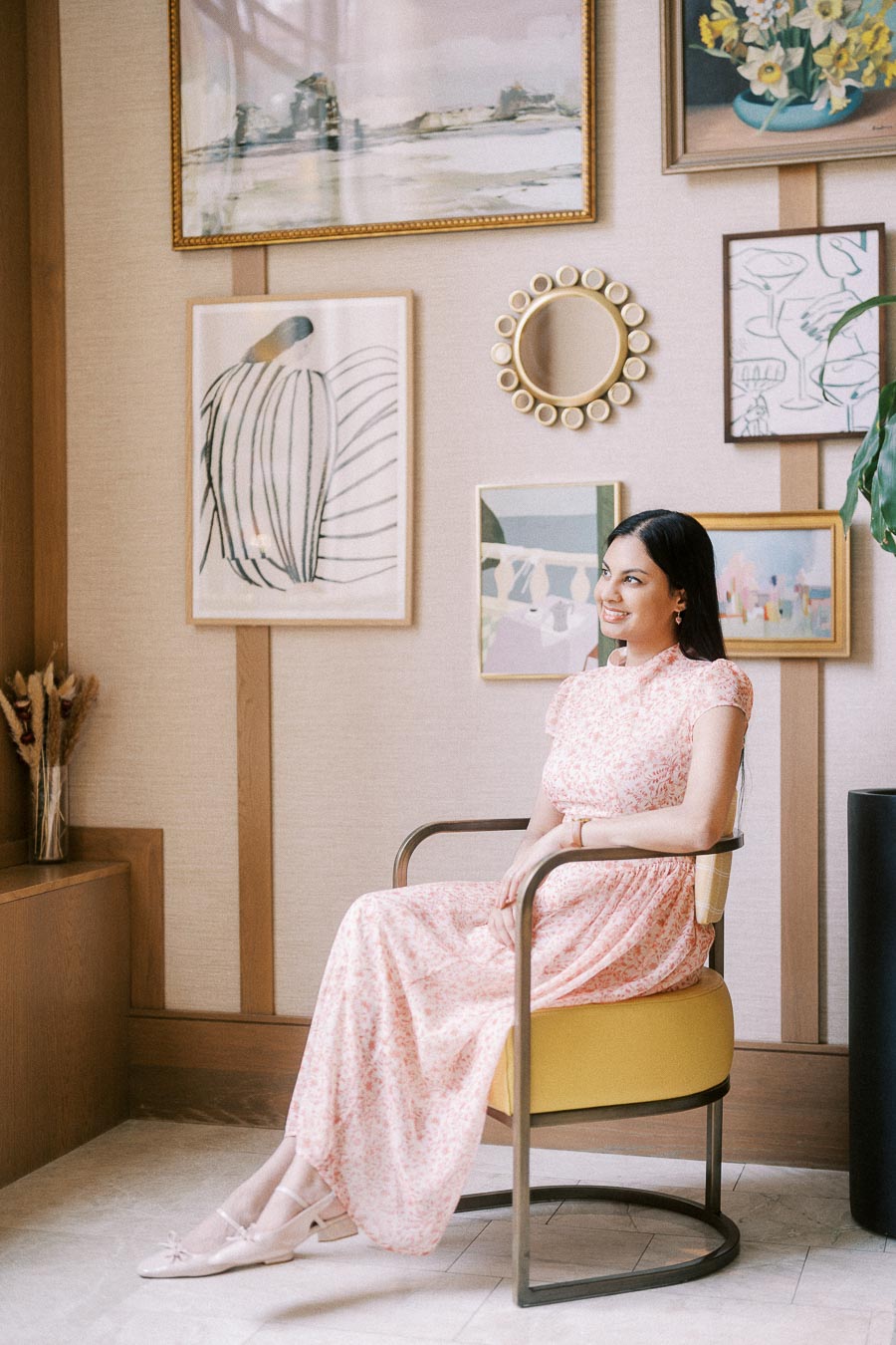 Becky seated in a pink floral dress beside a gallery wall of framed art