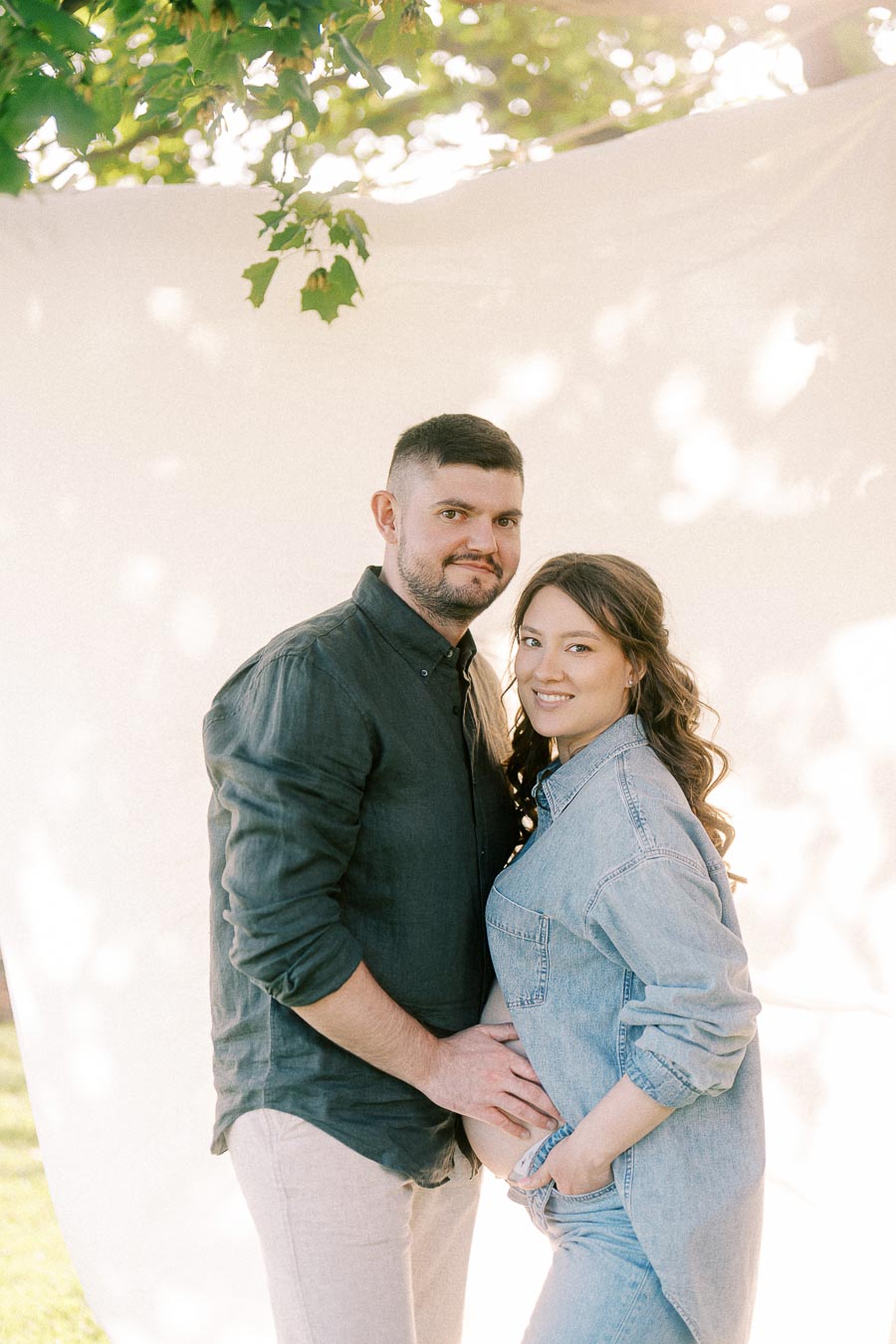 Expectant couple posing under leafy branches at The Green Acres