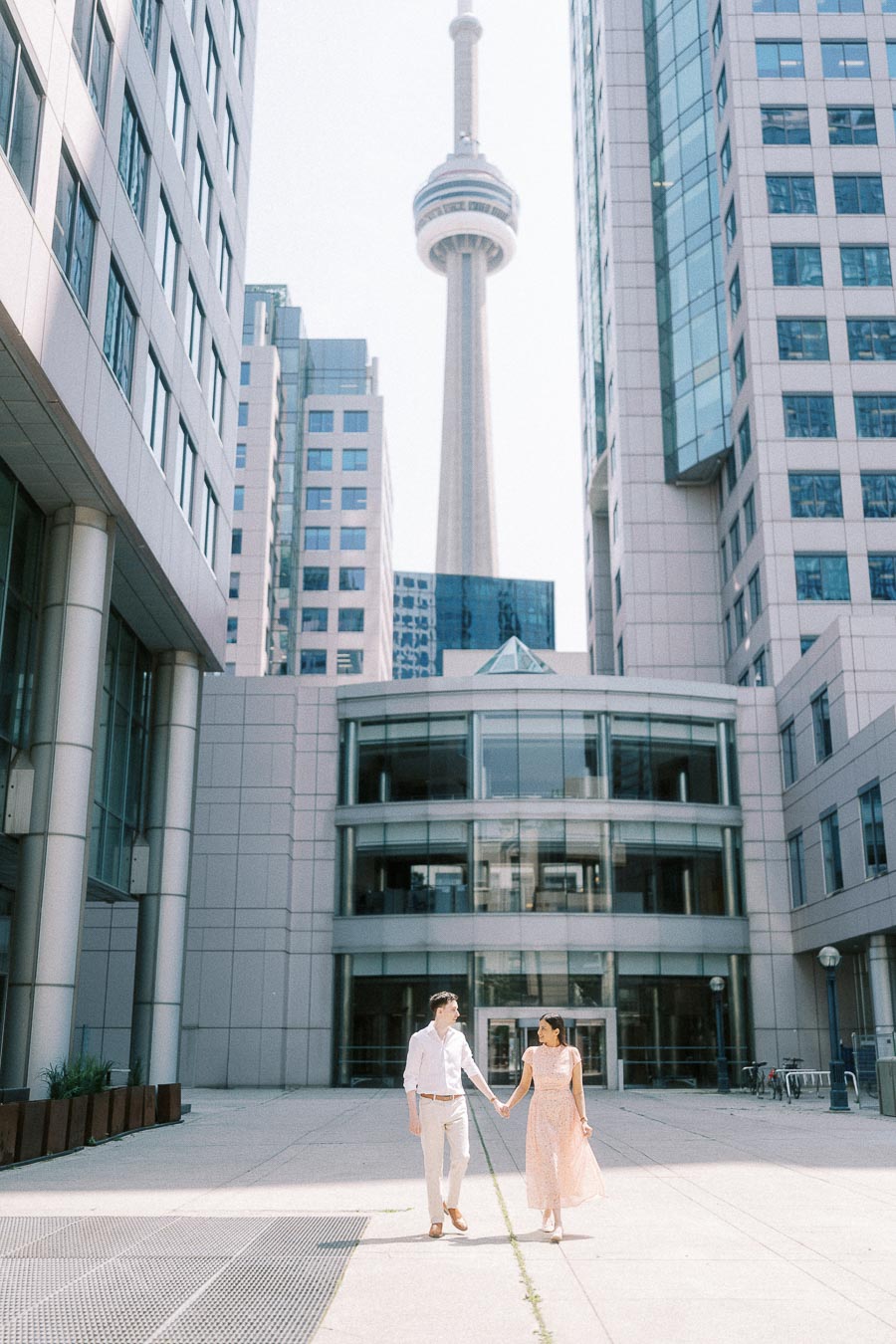 Couple holding hands beneath the CN Tower between downtown Toronto buildings