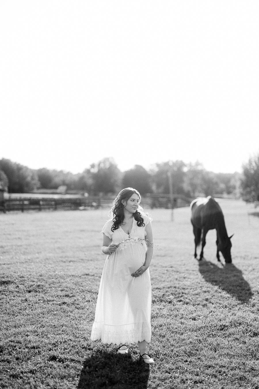 Pregnant woman in a white dress standing in a pasture with a grazing horse