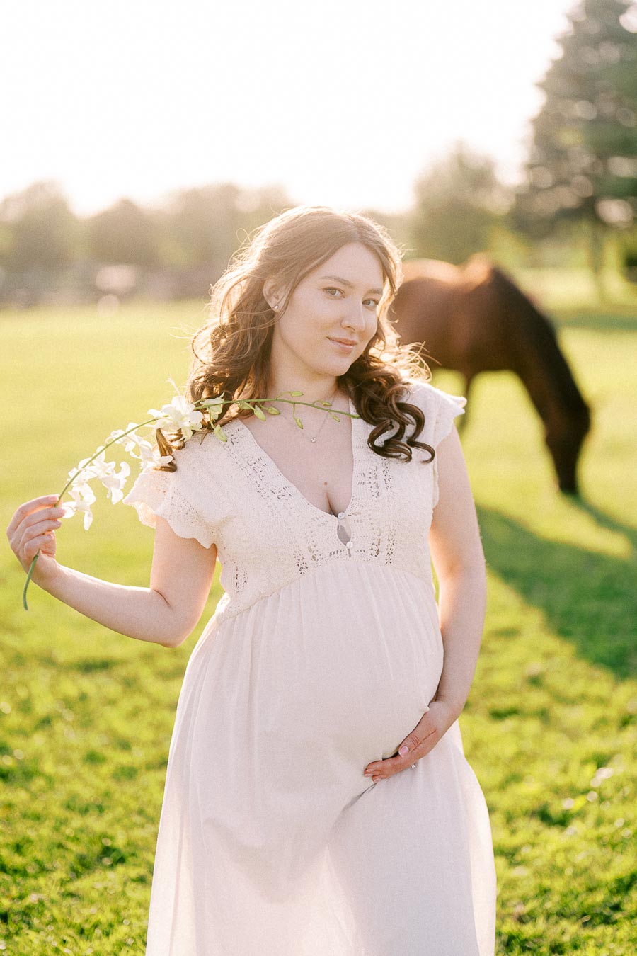 Pregnant woman in white dress holding flowers with a horse grazing behind her