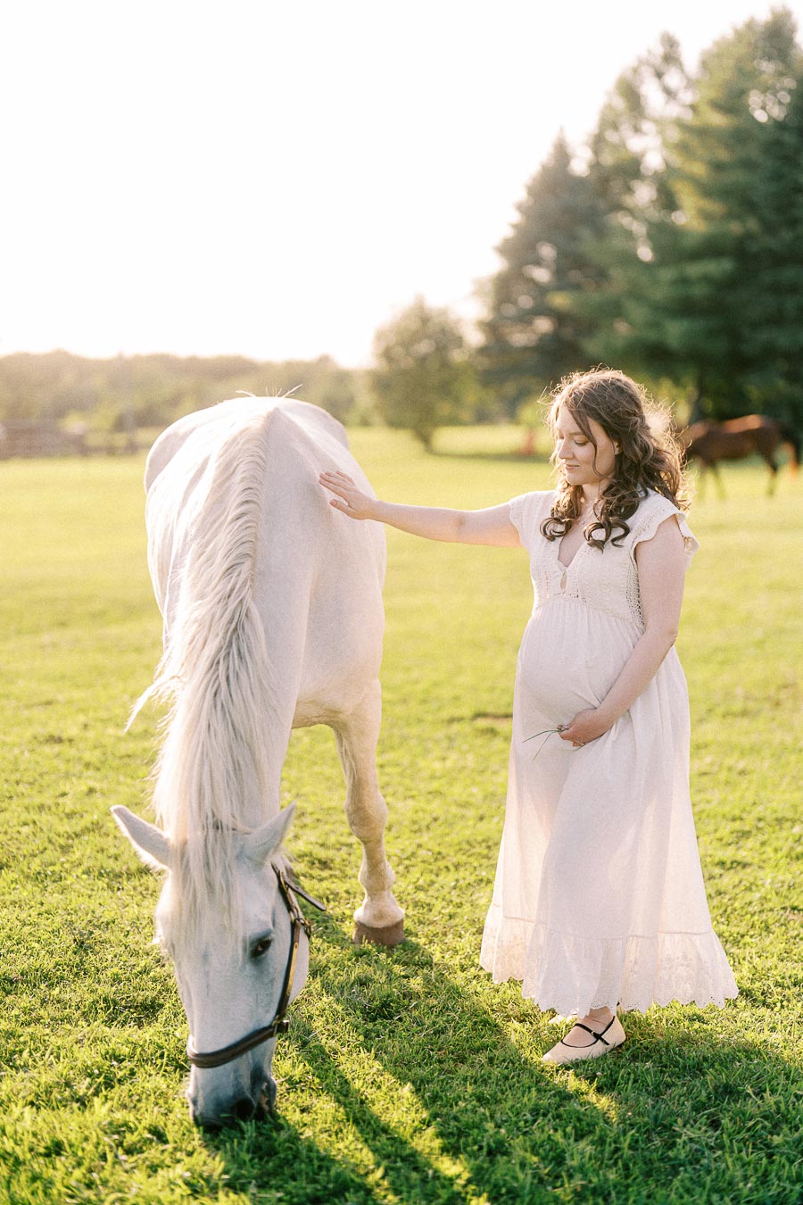 Pregnant woman in a white dress standing beside a grazing white horse