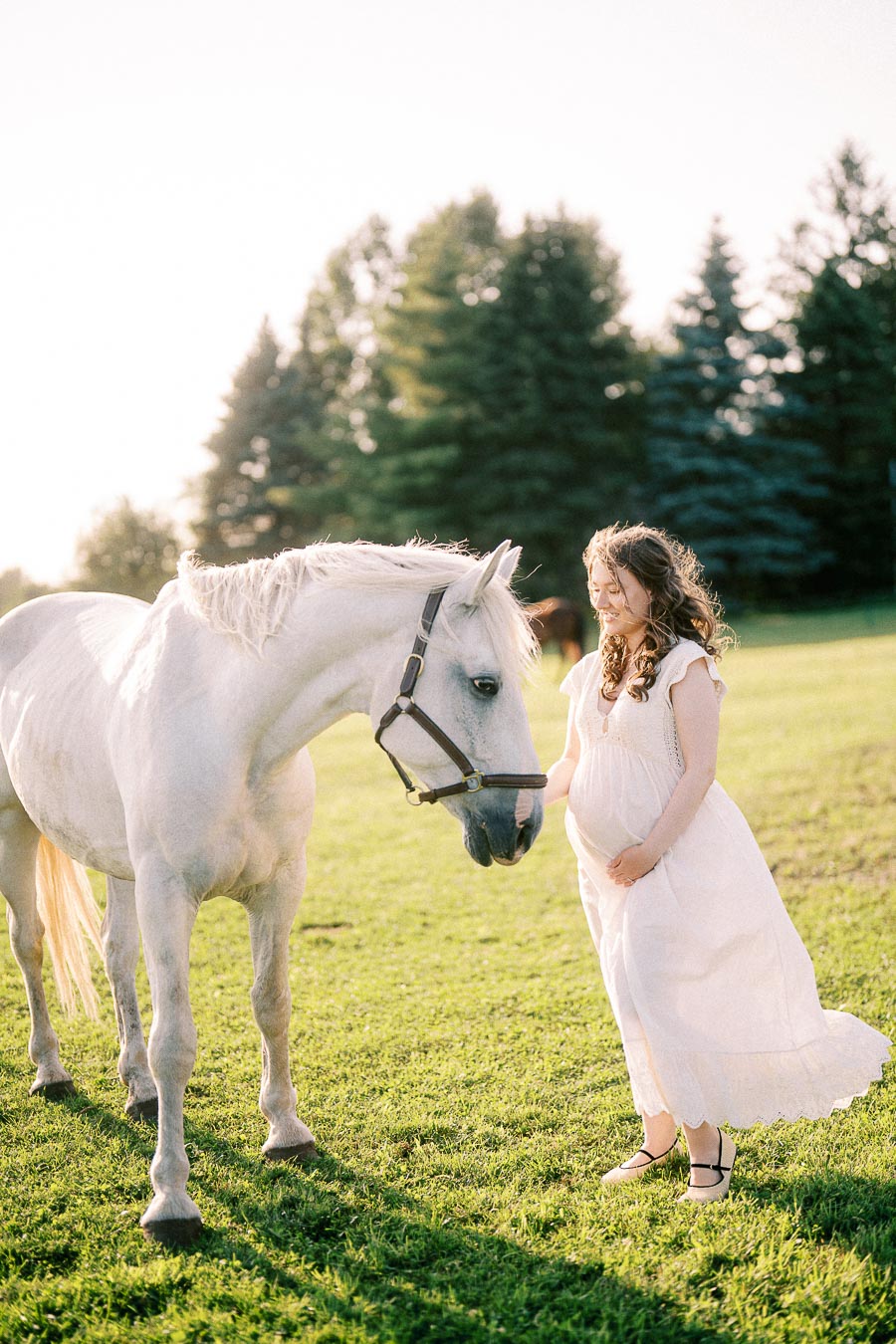 Pregnant woman in a white dress standing beside a white horse in a pasture