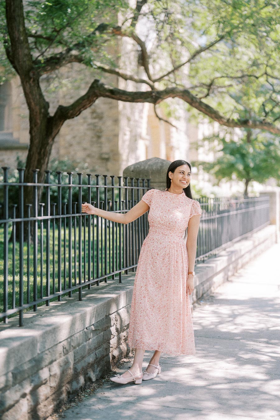 Becky in a pink floral dress beside an iron fence on a sunny Toronto sidewalk