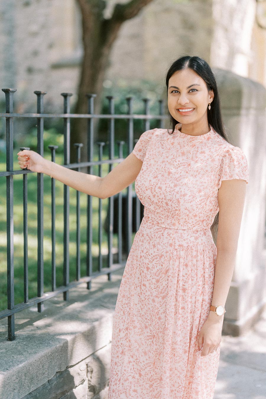 Becky smiling beside a black iron fence in a pale pink dress