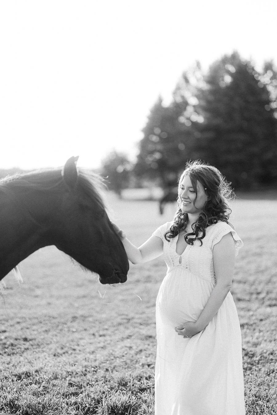 Pregnant woman in a white dress gently touching a horse in a field