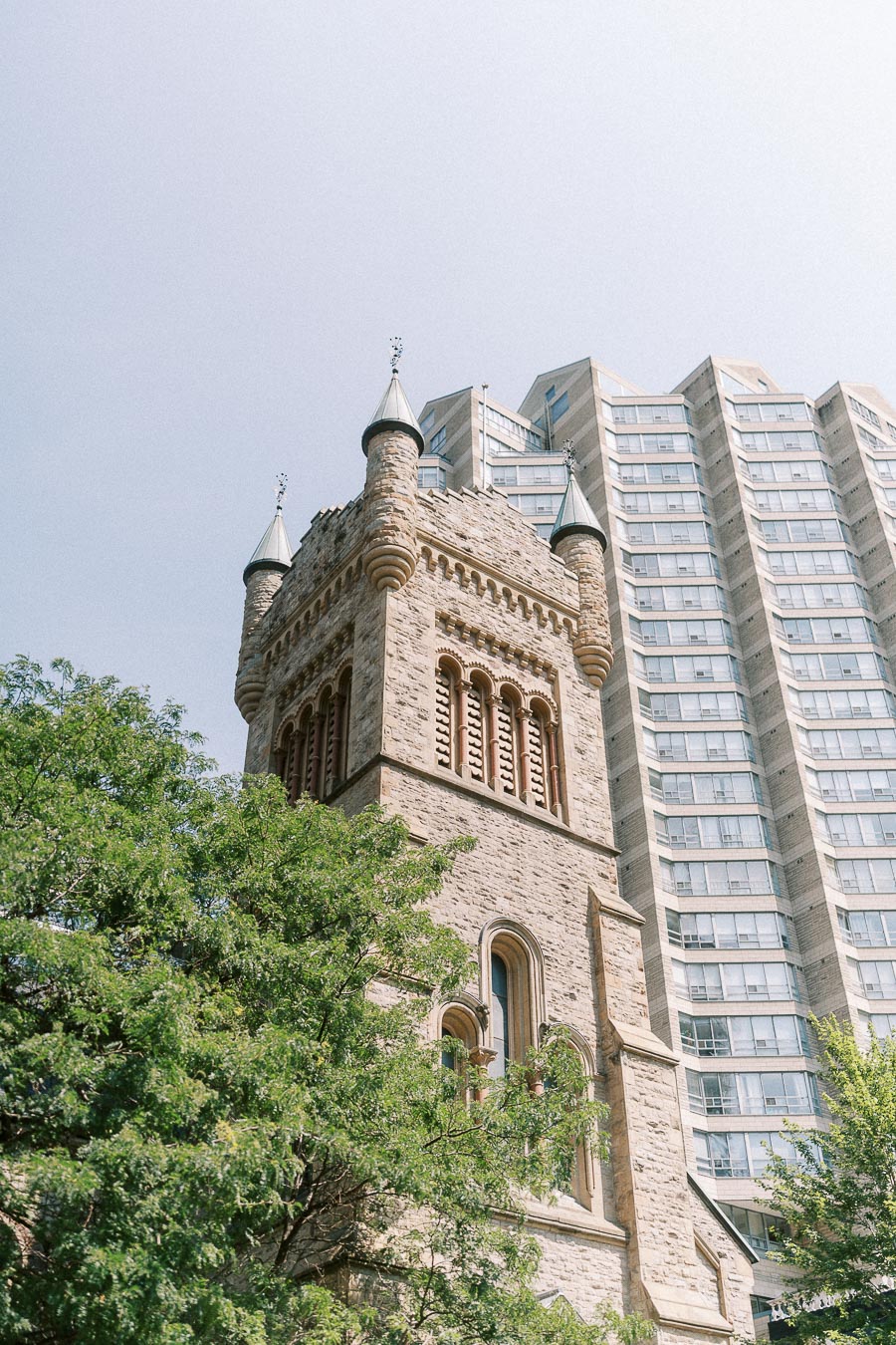 Historic stone church tower framed by trees and modern Toronto high-rise