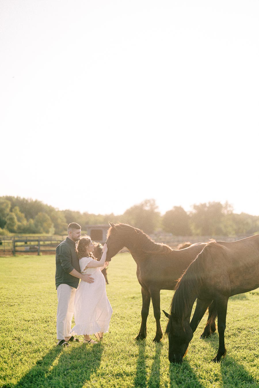 Expectant couple stands with two horses in a grassy field at The Green Acres