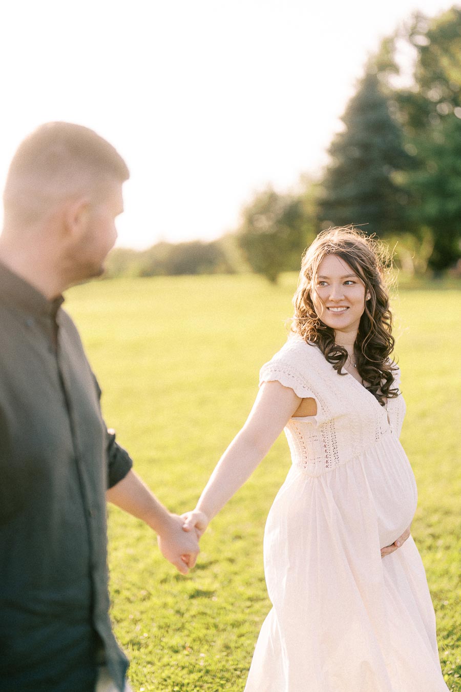 Expectant couple holding hands in a sunny field at The Green Acres