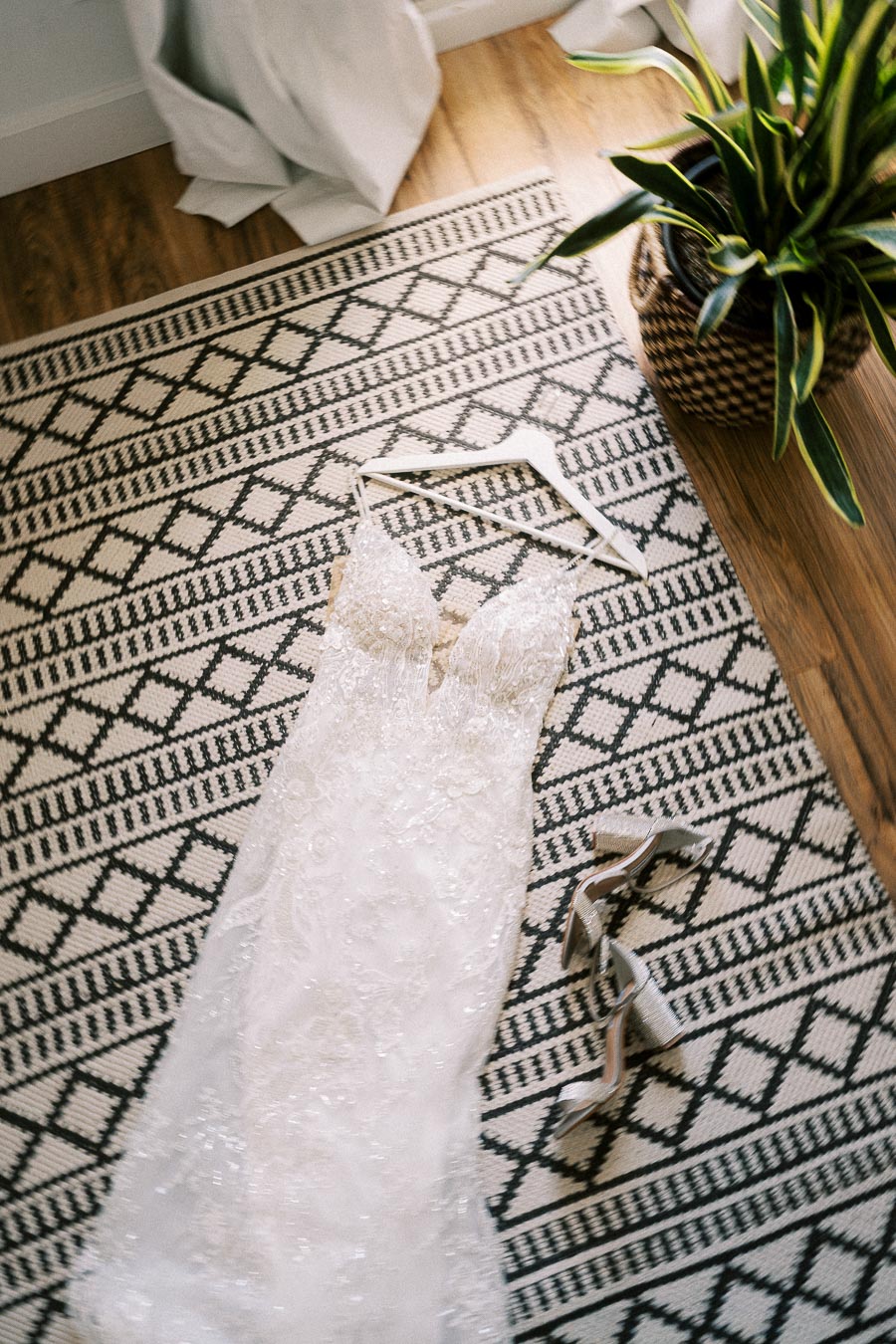 Beaded white wedding dress on a hanger with silver heels on a patterned rug beside a potted plant.