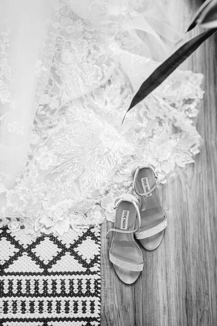 Black-and-white close-up of Steve Madden bridal heels beside a lace wedding dress on a wooden floor.