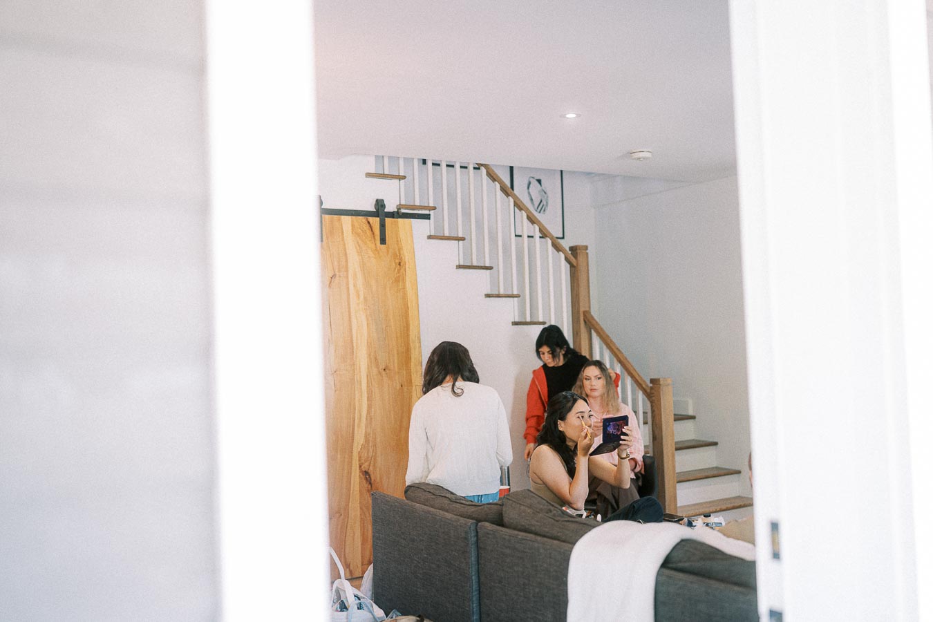 Four women sit and stand in a bright room by a staircase as one applies makeup using a compact mirror.
