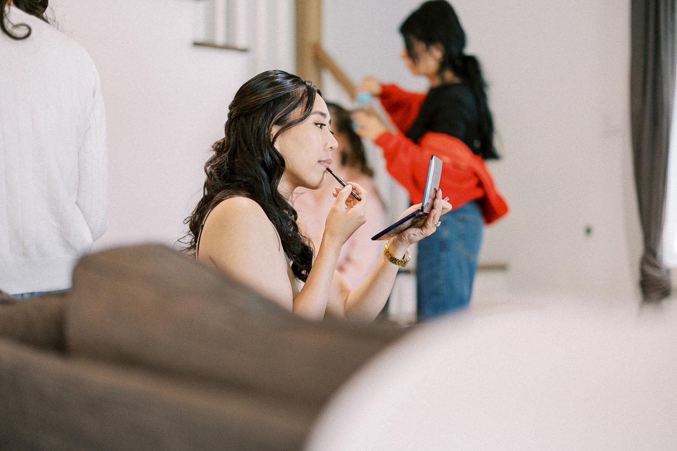 Bride applies lipstick while holding a mirror as attendants get ready in the background