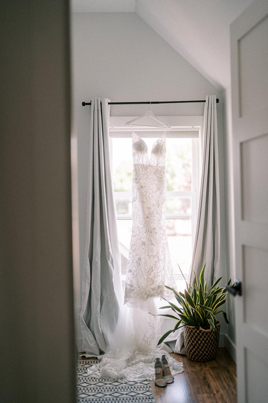 Lace wedding dress hanging in a bright window between curtains, with bridal shoes and a potted plant below.